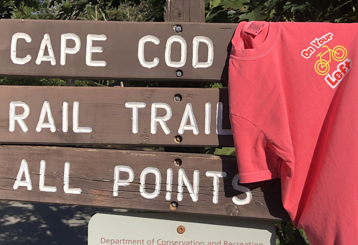 Cape Cod Rail Trail donation t-shirt hanging on a rustic wooden park sign to support trail projects.