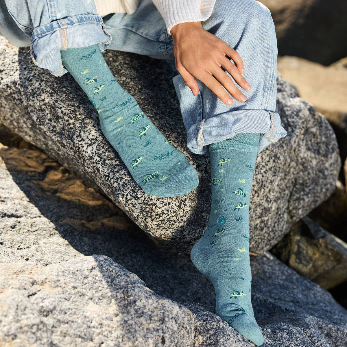 A person wearing organic cotton sea turtle protection socks while sitting on a coastal rock at a sandy beach.