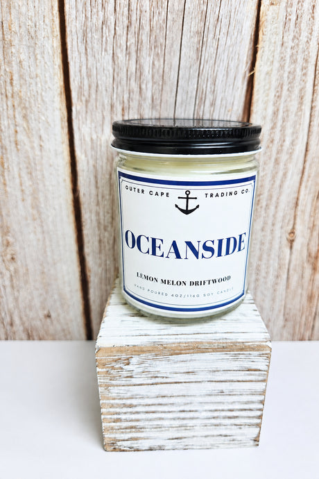 A glass jar candle with a black lid and a white label reading 'OCEANSIDE' and 'Lemon Melon Driftwood' by Outer Cape Trading Co, displayed on a distressed white block against a rustic wood plank background.