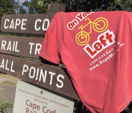 Cape Cod Rail Trail t-shirt hanging on a wooden bike trail sign, with proceeds from the shirt supporting trail maintenance.