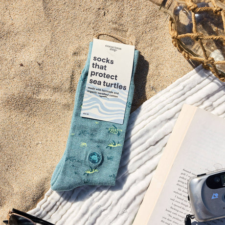 A pair of organic cotton sea turtle protection socks in eco-friendly retail packaging, beautifully displayed on the sand at a scenic beach.